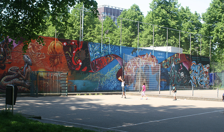 Children play basketball on an urban courtyard surrounded by colorful graffiti and plenty of trees.